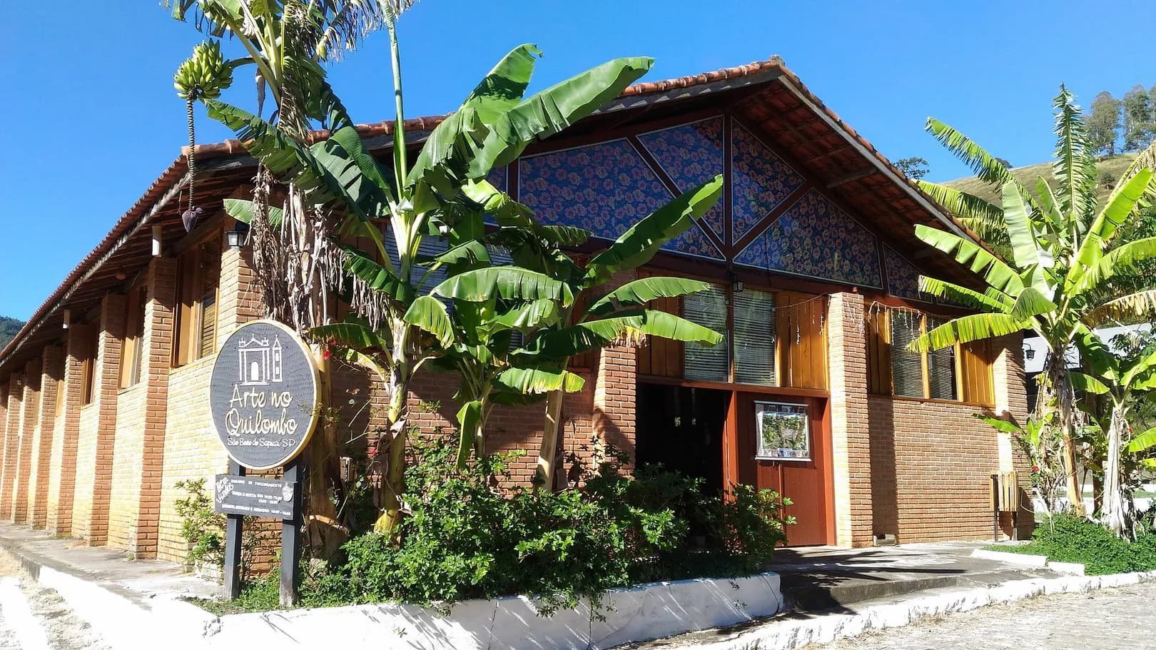 Fachada de tijolo e madeira do Arte no Quilombo, com telhado de telha, bananeiras na entrada e o morro ao fundo