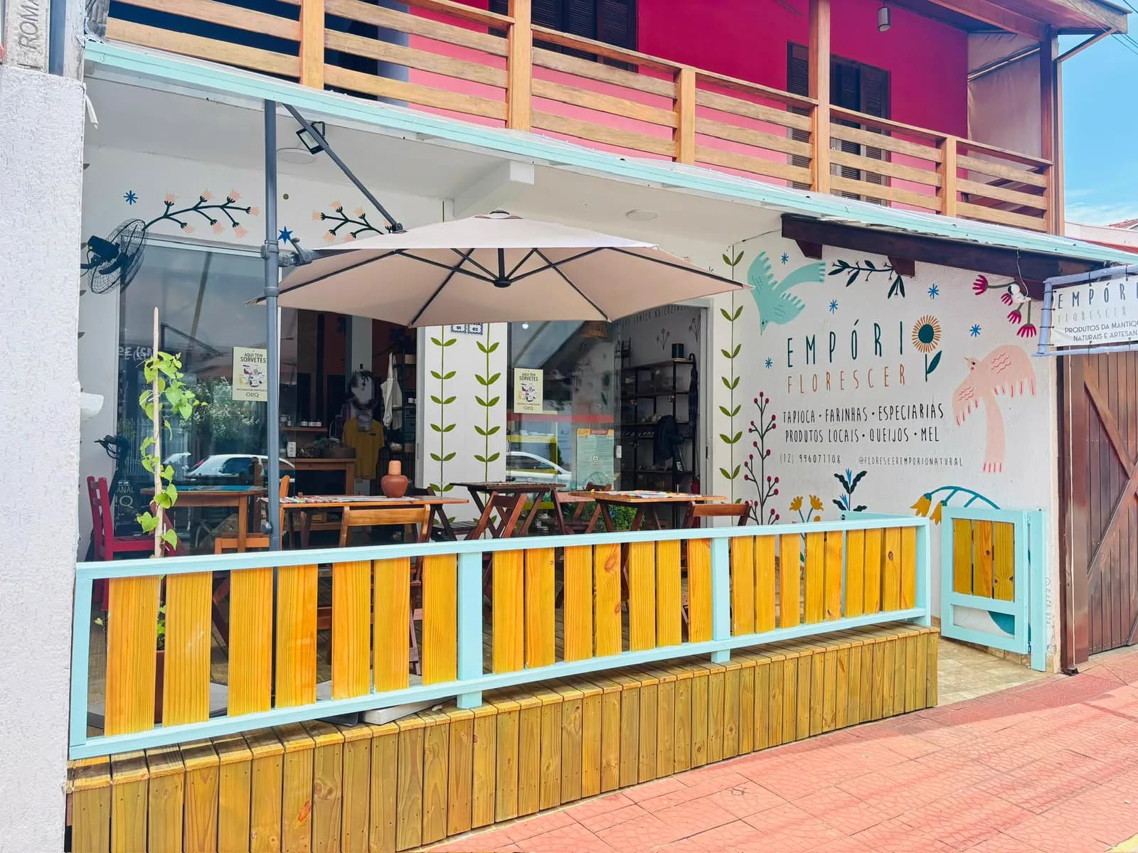 Fachada com deck de madeira, mesas sob guarda-sol e mural de flores e pássaros com o letreiro Empório Florescer