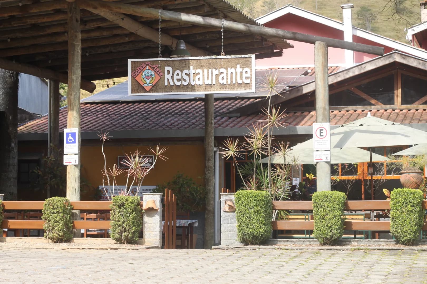 Entrada do restaurante Sabor com Arte, com a placa suspensa sob o pergolado de eucalipto, o telhado de telha e o morro coberto de mata atrás